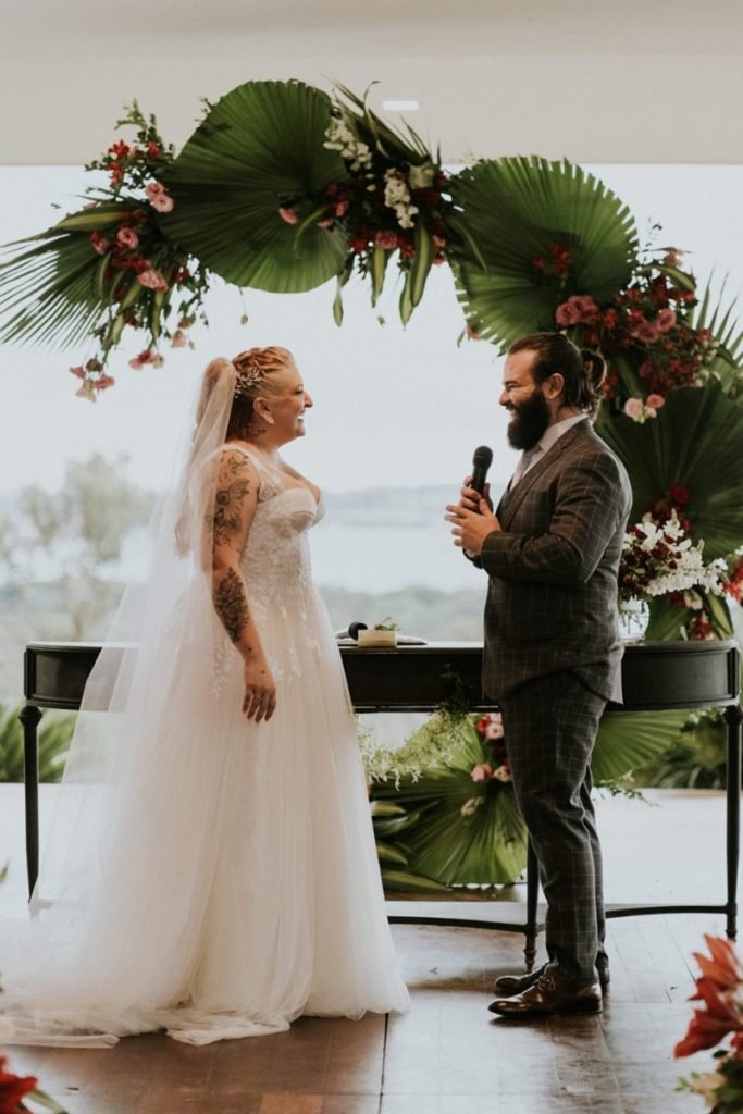 Pastel com caldo de cana: casal comemora união em pastelaria no DF - Veja fotos 7 Noivo segura microfone de frente para a noiva, ambos envoltos em arco de plantas