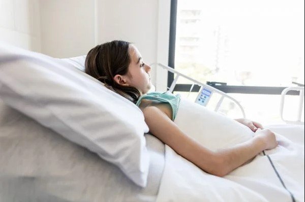 Na imagem colorida, uma crinaça com cabelos longos e pretos está deitada sobre uma cama de hospital