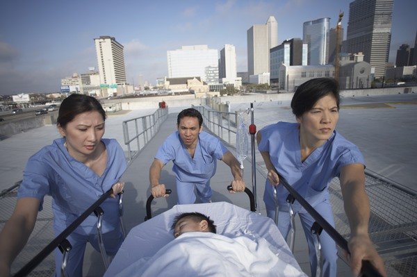Na imagem colorida, pessoas com roupas semelhante a de hospitais correm segurando uma maca com criança deitada