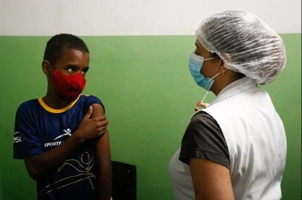 Na imagem colorida, uma criança está de frentre para uma enfermeira com uma seringa nas mãos