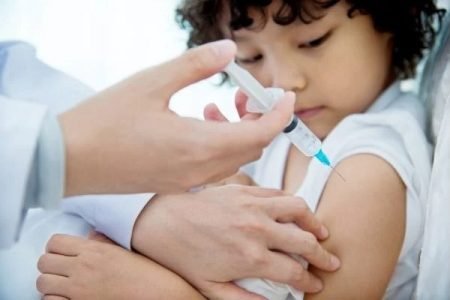 Na imagem colorida, uma criança está sentada enquanto alguém aplica uma vacina no braço dela