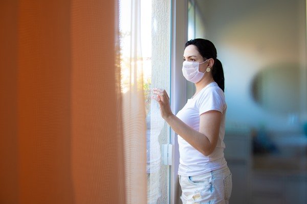 Na imagem colorida, uma mulher olha pela janela. Ela está em pé e usa mascara