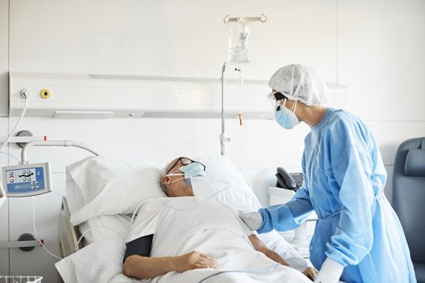 Na imagem colorida, uma pessoa está deitada em uma maca de hospital e outra pessoa de azul está com as mãos no braço dela.  Mascaras todos usam
