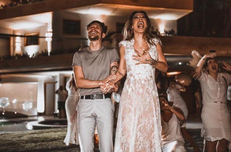 Na imagem colorida, um homem e uma mulher estão posicionados no centro. Ela usa vestido Branco de noiva e ele usa terno escuro com estampa dourada. Eles sorriem
