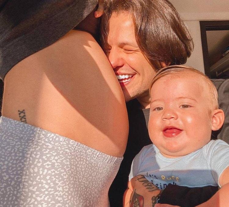 Na imagem colorida, há três pessoas posicionadas no centro. A barriga de uma delas aparece do lado esquerdo e do lado direito um homem beija a barriga enquanto segura um bebê no colo