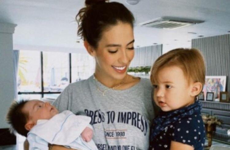 Na imagem colorida, uma mulher segura dois bebês no colo. Ela usa blusa cinza e está com os cabelos presos
