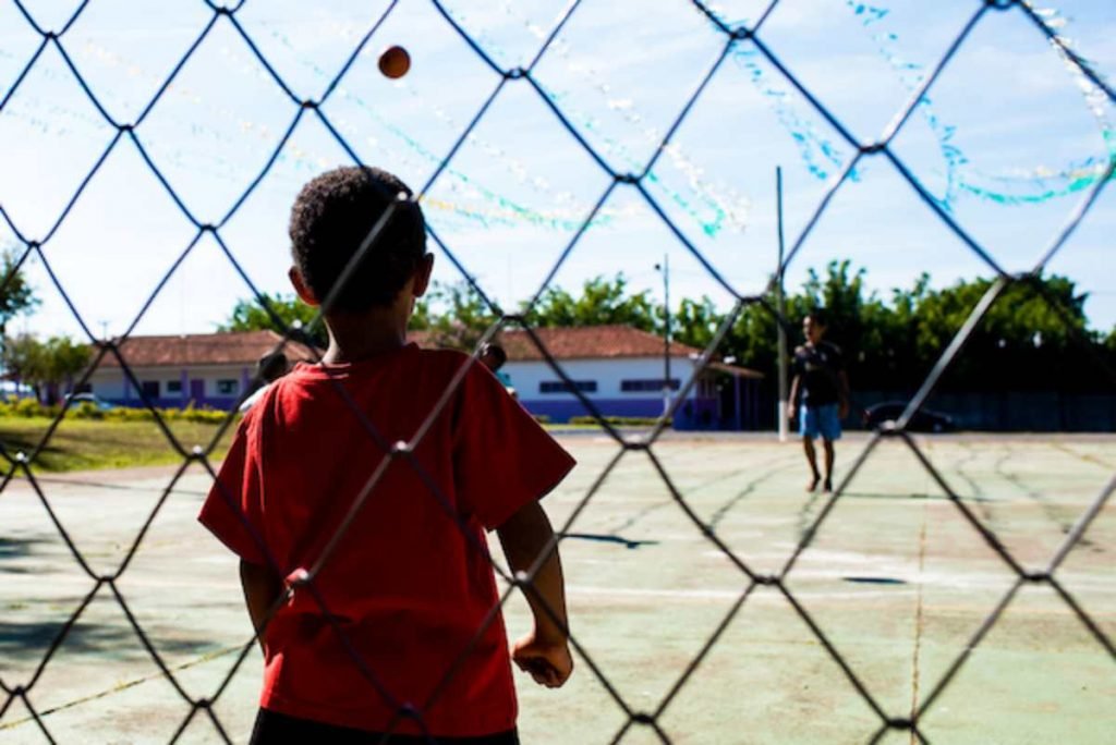 criança negra com camiseta vermelha atrás de uma grade, ao que parece ser de uma quadra esportiva