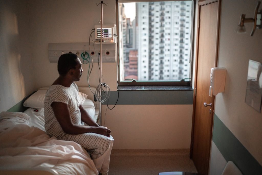 Fotografia colorida. homem sentado em cama de hospital olha pela janela