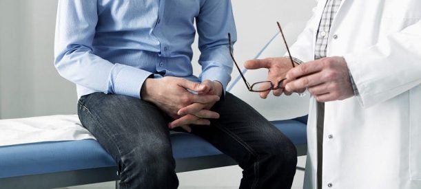 Fotografia colorida. Paciente sentado em maca com as mãos cruzadas conversa com médico que segura óculos nas mãos