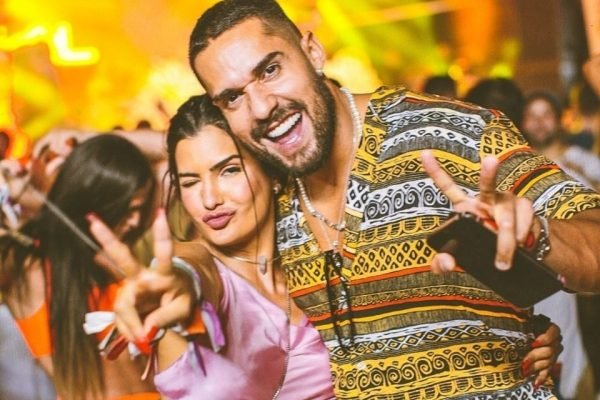 Mari e Bil posam abraçados para foto. Os dois levantam dois dedos de cada mão em show-Metrópoles