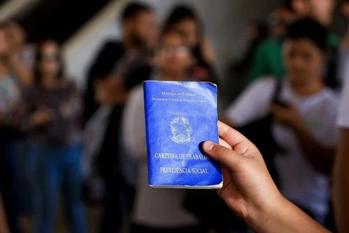 Mão segurando carteira de trabalho e pessoas passando ao fundo, desfocadas