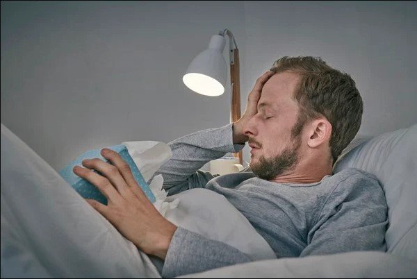 Na imagem colorida, um homem está deitado em uma cama com as mãos na cabeça
