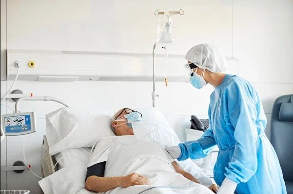 In the color picture, a person is lying in a hospital wheelchair, and another person in blue has his hands on his arm.  everyone wears masks