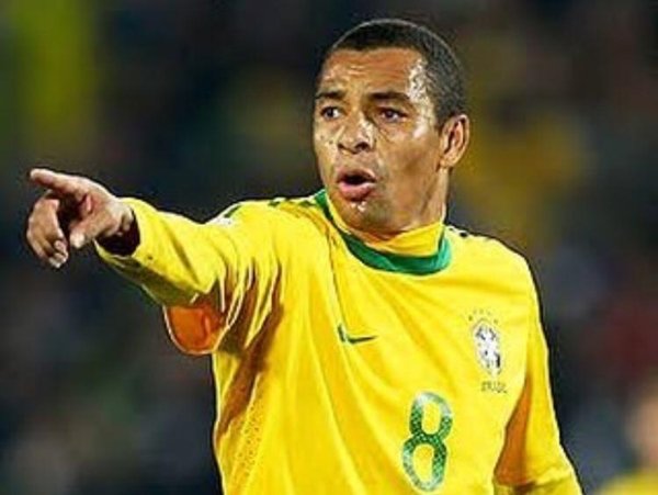 Na imagem colorida, um homem está centralizado no centro. Ele usa camisa do Brasil e está em um campo de futebol