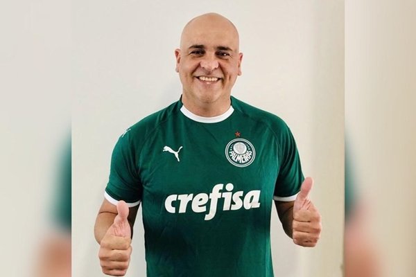 Na imagem colorida, um homem está centralizado no centro. Ele usa camisa de time de futebol brasileiro e faz sinal de joia com as mãos