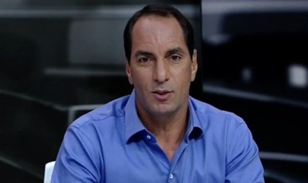 Na imagem colorida, um homem está posicionado no centro. Ele usa camiseta azul e olha atentamente para a câmera