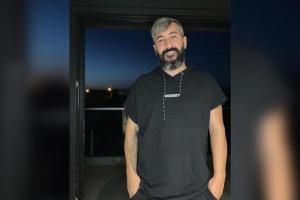 Na imagem colorida, um homem está posicionado no centro. Ele usa camiseta e calça nas cores preta e sorri para a câmera