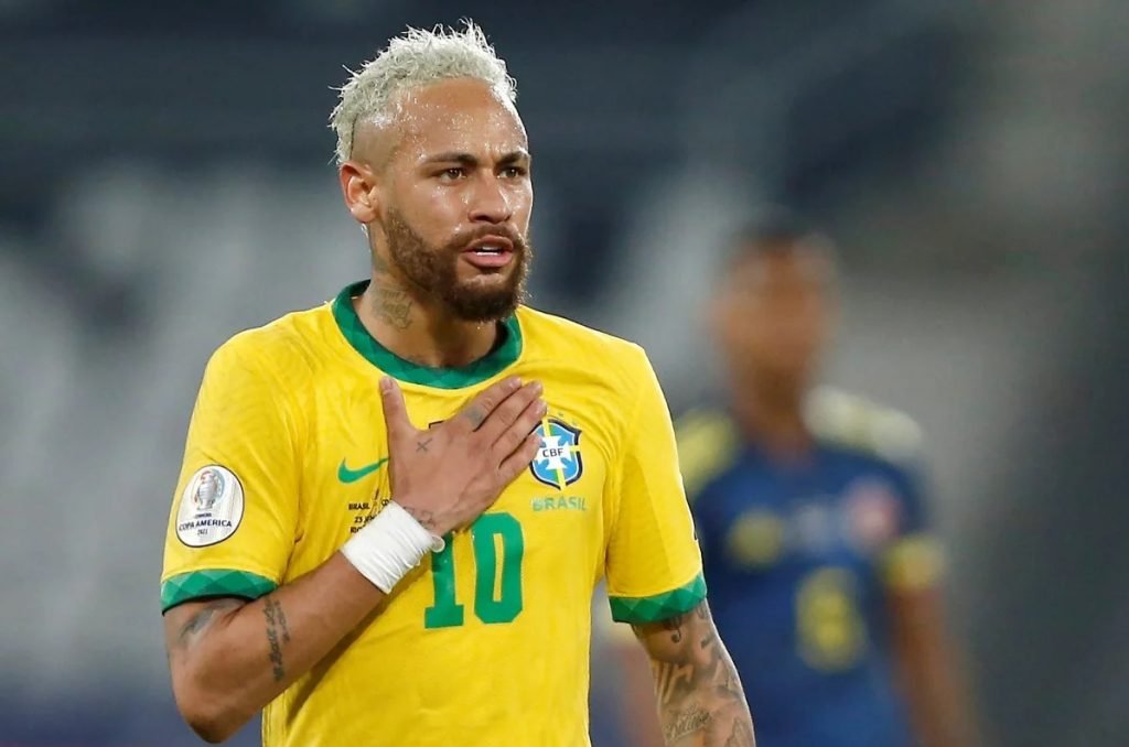 Na imagem colorida, um homem está centralizado no centro. Ele usa camisa do Brasil e está em um campo de futebol