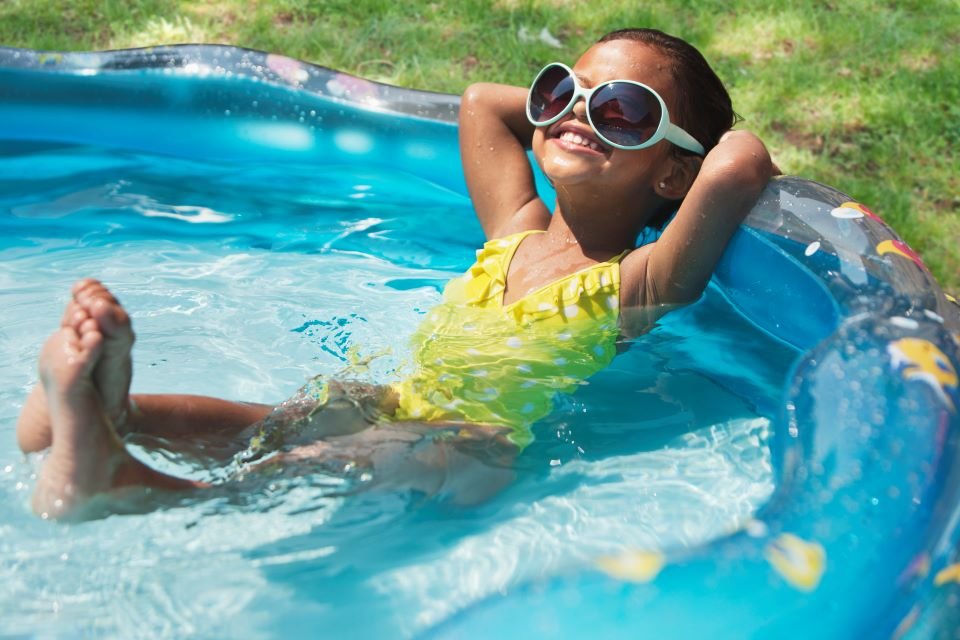 Criança de maiô verde e óculos de sol em piscina de plástico no sol - Metrópoles