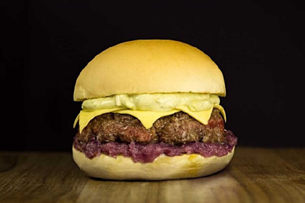 Na foto temos um hamburguer com carne e queijo e um molho roxo
