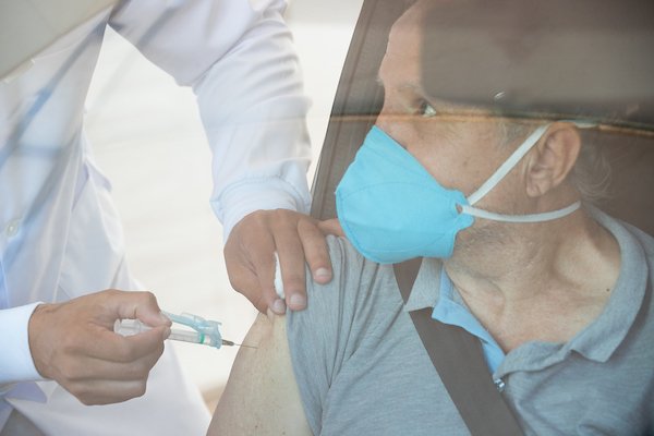 Pessoa de máscara dentro do carro e mão aplicando vacina no braço