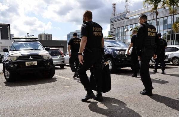 Na imagem colorida, pessoas estão posicionadas à direita. Eles usam roupa preta com o nome da Policia Federal. Eles andam por um estacionamento de carros