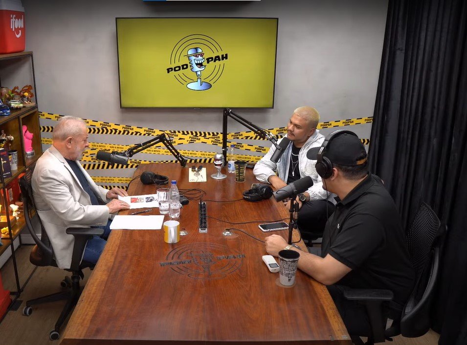 Ex-presidente Lula no podcast do YouTube PodPah. Ele está na frente dos apresentadores Igão e Mítico, numa mesa com três microfones e um telão ao fundo com a logo do podcast - Metrópoles