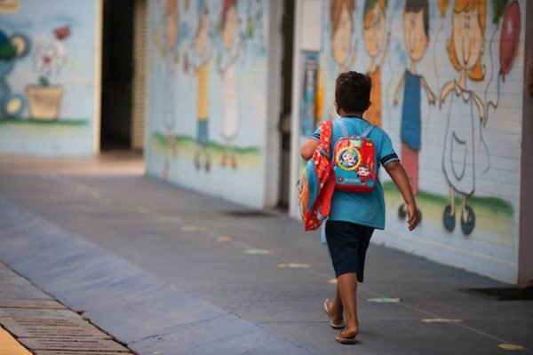 Menino de uniforme azul, de costas, com mochila vermelha, caminhando em escola