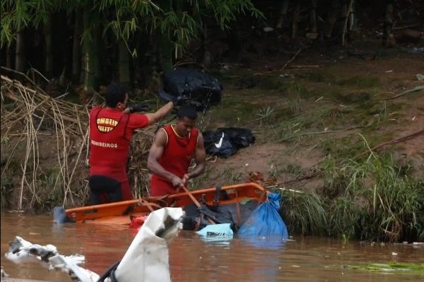 Na imagem colorida, homens se posicionam à esquerda. Eles estão dentro de um rio com lama e objetos e usam roupa vermelha