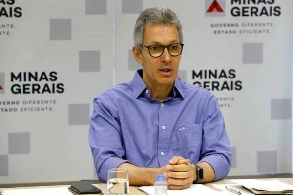 Na imagem colorida, um homem está posicionado no centro. Ele usa camiseta zul, usa oculos e olha atentamente para frente