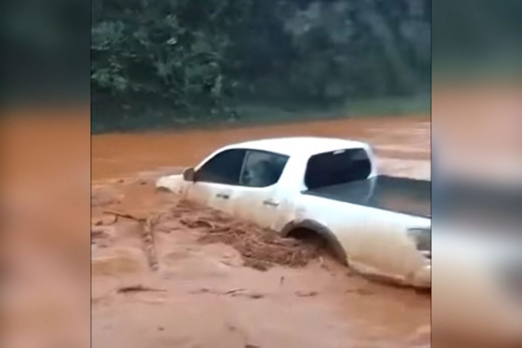Na imagem colorida, uma caminhonete branca está à direita. Ela é branca e está parcialmente submersa em lama