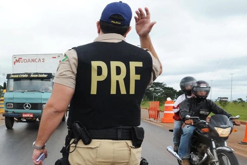 Na imagem colorida, um homem de costas com o colete da PRF está posicionado no centro. Do lado direita há uma moto com duas pessoas em cima e do lado esquerdo há um caminhão branco com azul