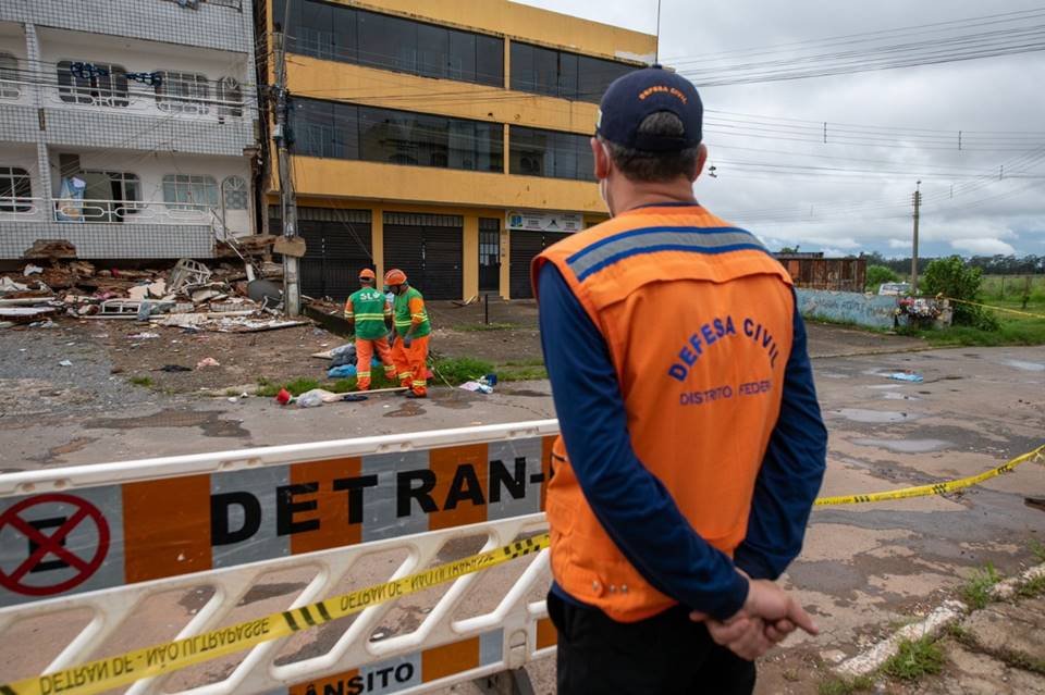 Homem com uniforme da Defesa Civil observa dois garis catando lixo