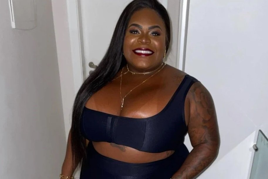Na imagem colorida, uma mulher está posicionada a direita.Ele usa roupas escuras, tem cabelos longos e sorri para a camera