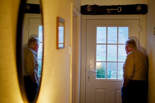 An elderly man standing by a door looking out through the window panes.