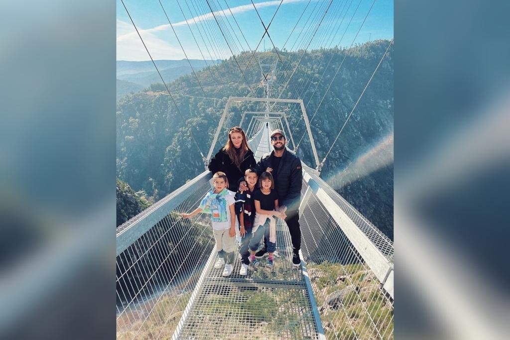 Na imagem colorida, pessoas estão posicionadas no centro. Elas estão em cima de uma ponte branca e sorriem para a camera