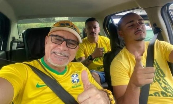 Na imagem colorida, três homens ocupam o centro da imagem. Eles estão dentro de um carro, usam camiseta do Brasil, fazem o símbolo de joia com as mãos e estão sorrindo para a camera
