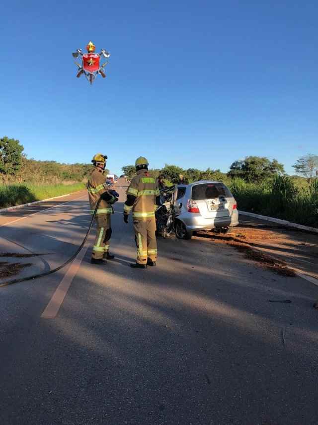 dois bombeiros vestidos de amarelo com mangueira apontada para um carro prata parcialmente destruído