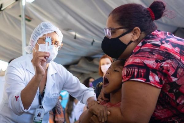 Mulher de máscara segurando menino de boca aberta e enfermeira mostrando agulha com vacina