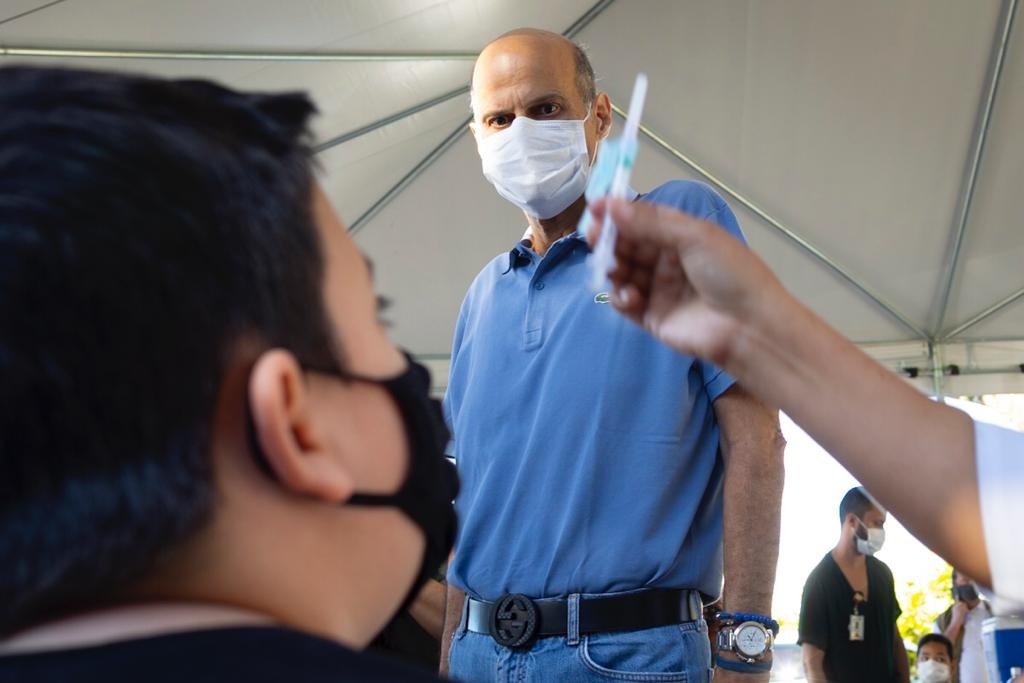 Rosto de menino com máscara olhando para agulha e homem de azul ao fundo