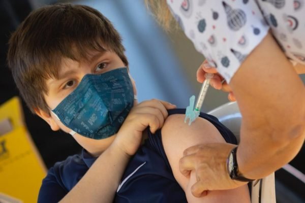 Menino com máscara azul, puxando a manga da camisa e mão aplicando vacina no braço dele