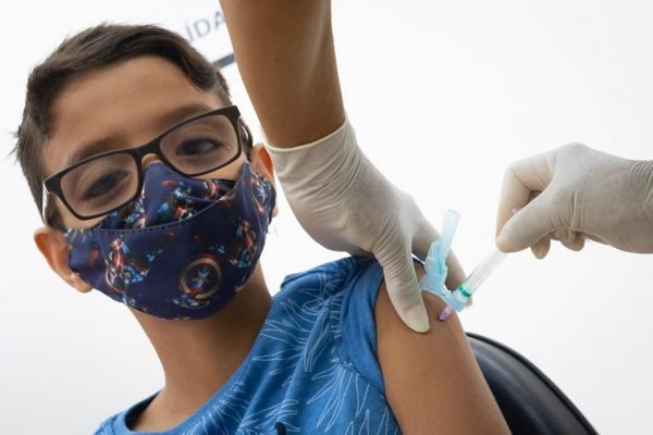Mão aplicando vacina em braço de menino de óculos e máscara