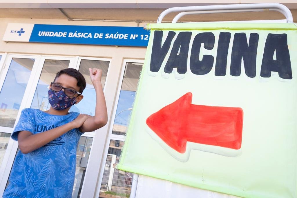 Menino de máscara e óculos com a mão para cima ao lado de placa escrito vacina com uma seta vermelha