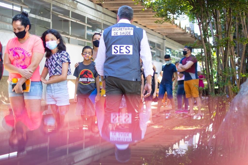 Homem com uniforme da SES de costas e fila de pessoas atrás