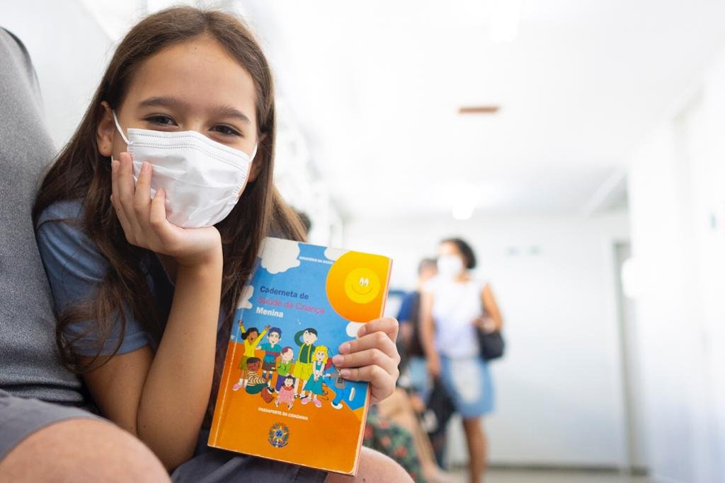 Menina com máscara branca e mão no queixo segurando livro com a outra mão