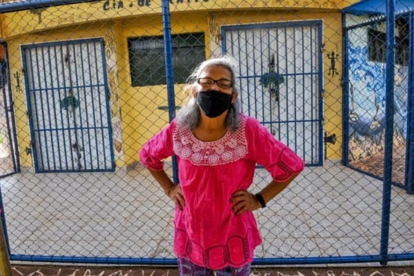 Idosa de blusa rosa e máscara preta com as mãos na cintura em frente a grade