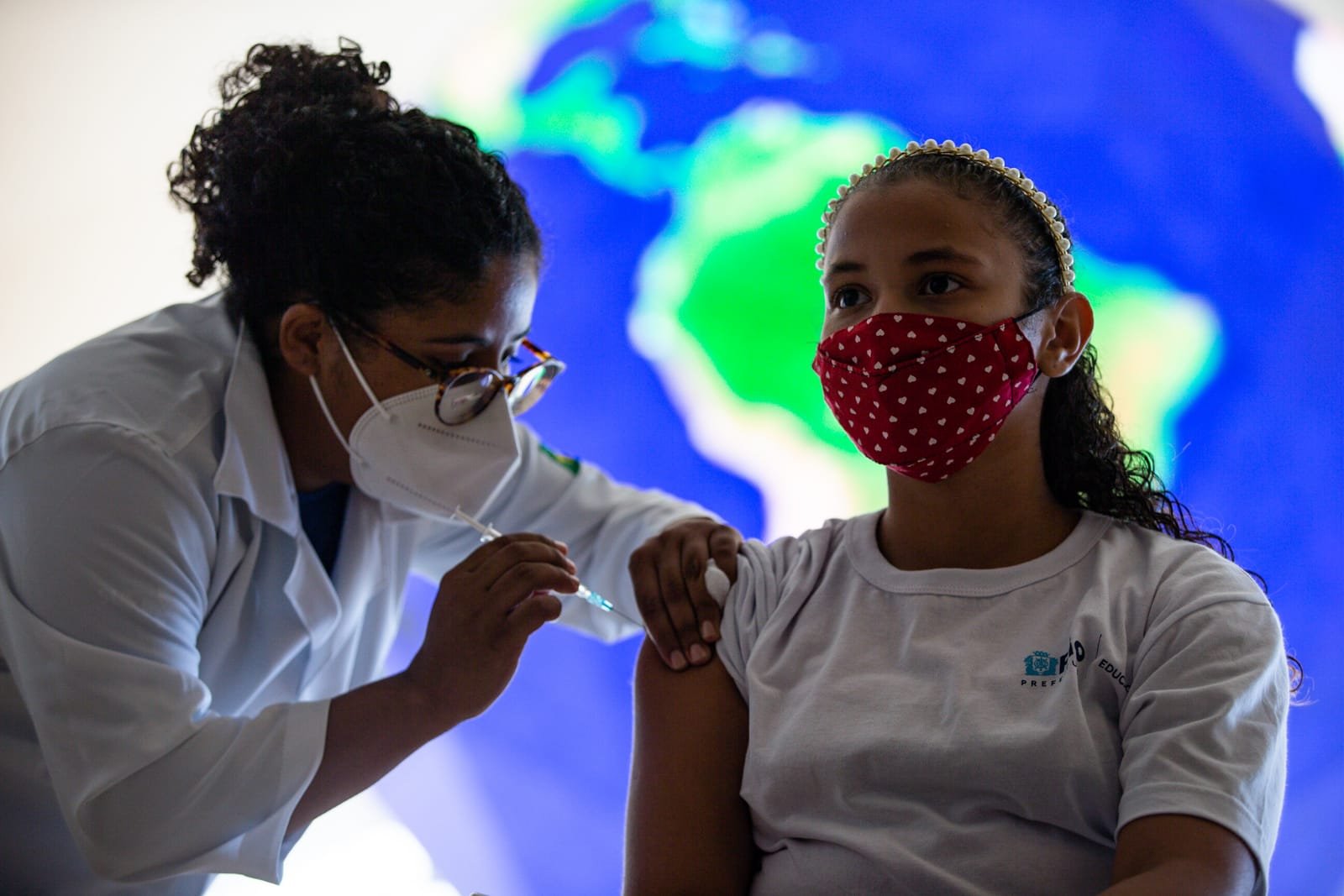 imagem colorida mostra criança com máscara de pano e camiseta branca sendo vacinada por enfermeira que usa roupa branca, máscara cirúrgica e óculos - Metrópoles