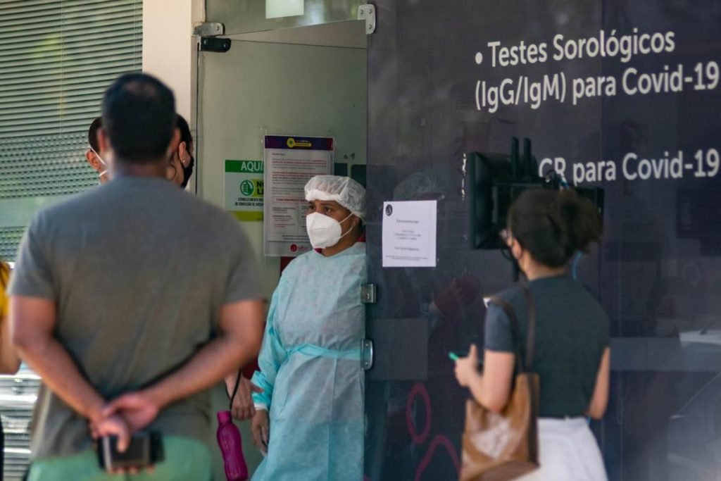 mulher com materiais de proteção - touca, roupa de proteção e máscara, conversando com dois homens que estão em uma longa fila