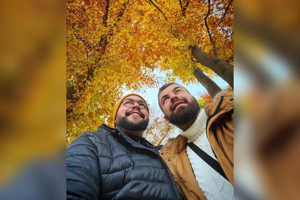 In the colorful image, two men are positioned in the center.  They are under the trees with yellow leaves, they wear cold jackets, headdresses, and smiley eyes on their foreheads