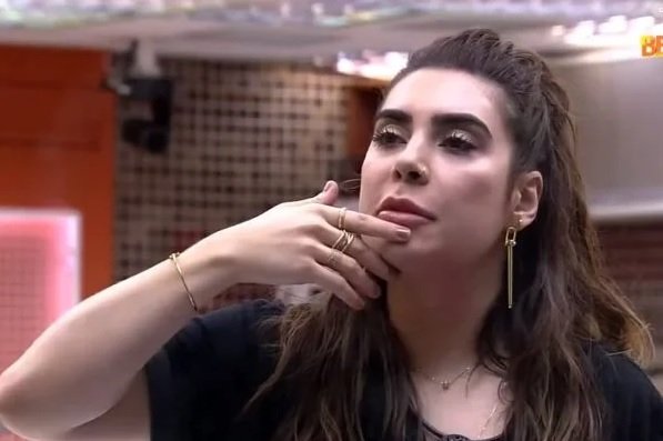 Na imagem colorida, uma mulher está posicionada no centro. Ela tem cabelos curtos e loiros, olhos castanhos e olha seriamente para a câmera